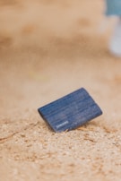 A blue portable Samsung solid state drive is partially buried in sand, with a blurred sandy background. The surface appears to be dusty, suggesting an outdoor environment.