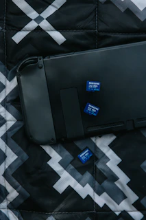 A black electronic device lies on a patterned fabric surface alongside three blue Samsung SD cards. The fabric has a geometric design in shades of black, white, and gray.