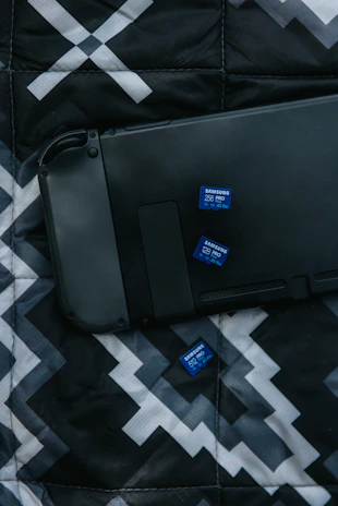 A black electronic device lies on a patterned fabric surface alongside three blue Samsung SD cards. The fabric has a geometric design in shades of black, white, and gray.
