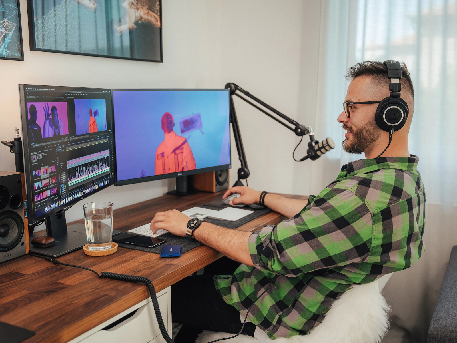 A creator wearing headphones edits a video on two monitors at a home desk with a boom microphone
