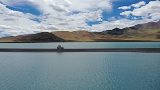 A vehicle is parked on a narrow strip of land surrounded by a calm, expansive body of turquoise water. Rolling hills and mountains rise in the background under a partly cloudy sky with varying shades of white and blue.