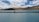 A vehicle is parked on a narrow strip of land surrounded by a calm, expansive body of turquoise water. Rolling hills and mountains rise in the background under a partly cloudy sky with varying shades of white and blue.