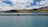 A vehicle is parked on a narrow strip of land surrounded by a calm, expansive body of turquoise water. Rolling hills and mountains rise in the background under a partly cloudy sky with varying shades of white and blue.