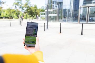 A person holding a smartphone with a financial application open on the screen, standing outside a modern glass building. The background shows a plaza with trees and pedestrians.