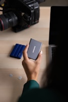A person holds a compact Samsung external hard drive near a laptop. Nearby, there are small memory cards and a camera with a large lens.