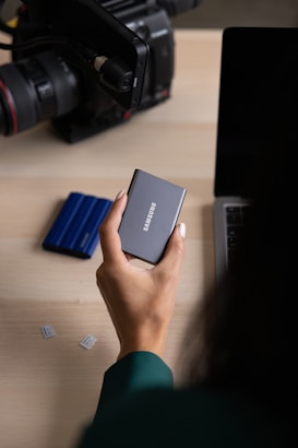 A person holds a compact Samsung external hard drive near a laptop. Nearby, there are small memory cards and a camera with a large lens.