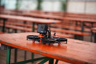 A small drone equipped with a camera is resting on a wooden table in an outdoor setting. The table has an orange surface with green metal legs. Several identical tables and benches are lined in the background, creating a pattern. The setting appears calm and peaceful, likely a park or an outdoor dining area.