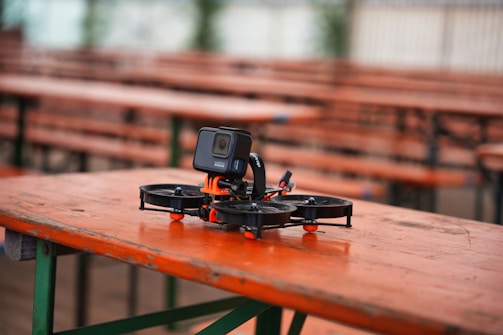 A small drone equipped with a camera is resting on a wooden table in an outdoor setting. The table has an orange surface with green metal legs. Several identical tables and benches are lined in the background, creating a pattern. The setting appears calm and peaceful, likely a park or an outdoor dining area.