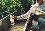 A person is filling a plastic bottle with water from an outdoor faucet, surrounded by a lush green background. The individual is wearing a light-colored hoodie and holding additional items under their arm.