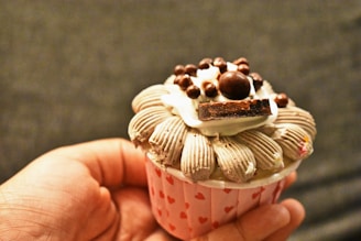 A hand is holding a cupcake with intricate frosting designs and various toppings, including chocolate pieces and small chocolate balls. The cupcake wrapper is decorated with small red hearts.