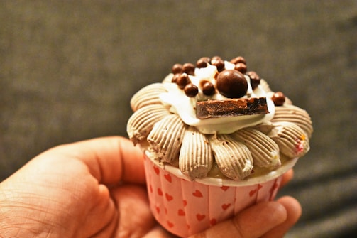 A hand is holding a cupcake with intricate frosting designs and various toppings, including chocolate pieces and small chocolate balls. The cupcake wrapper is decorated with small red hearts.