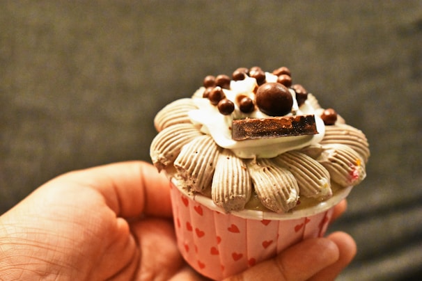 A hand is holding a cupcake with intricate frosting designs and various toppings, including chocolate pieces and small chocolate balls. The cupcake wrapper is decorated with small red hearts.