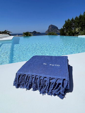 A luxurious swimming pool with clear blue water reflects the bright sky. A large, blue towel with the words 'El Patio' is elegantly folded on the poolside. In the background, a rocky island is visible across the sea, surrounded by lush greenery.