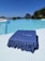 A luxurious swimming pool with clear blue water reflects the bright sky. A large, blue towel with the words 'El Patio' is elegantly folded on the poolside. In the background, a rocky island is visible across the sea, surrounded by lush greenery.