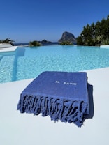 A luxurious swimming pool with clear blue water reflects the bright sky. A large, blue towel with the words 'El Patio' is elegantly folded on the poolside. In the background, a rocky island is visible across the sea, surrounded by lush greenery.