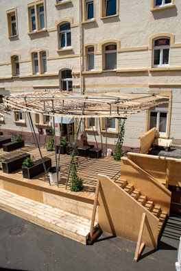 A wooden deck with a partially constructed pergola stands in front of a multi-story residential building with cream-colored walls and windows with shutters. Potted plants line the perimeter of the deck, and wooden steps lead up to the platform. Beneath the pergola framework, additional structural support is visible.