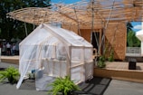 A small greenhouse structure with a transparent covering stands in the foreground, surrounded by lush green plants. Behind it is a modern wooden building with a distinctive slatted roof design, supported by metal beams. The area features a wooden deck and people are seen in the background, suggesting an outdoor event or exhibition.