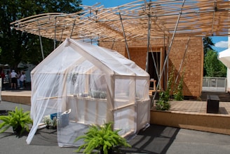 A small greenhouse structure with a transparent covering stands in the foreground, surrounded by lush green plants. Behind it is a modern wooden building with a distinctive slatted roof design, supported by metal beams. The area features a wooden deck and people are seen in the background, suggesting an outdoor event or exhibition.
