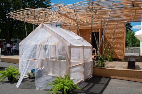 A small greenhouse structure with a transparent covering stands in the foreground, surrounded by lush green plants. Behind it is a modern wooden building with a distinctive slatted roof design, supported by metal beams. The area features a wooden deck and people are seen in the background, suggesting an outdoor event or exhibition.