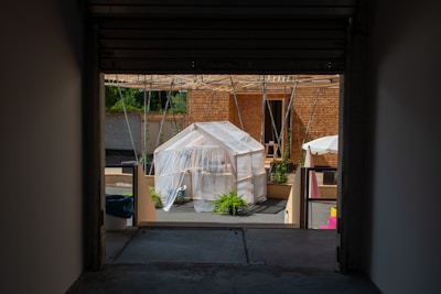 A small structure covered with translucent fabric is situated in a courtyard area with various plants around. The structure has a pitched roof and an open entrance. Behind it, there are wooden panels and vertical supports possibly for shade. There is also a white umbrella to the right and other elements like buckets and potted plants.