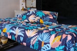 A bed is adorned with a vibrant and colorful floral-patterned bedding set. The design features an array of tropical leaves and abstract shapes in shades of blue, pink, yellow, and orange against a dark background. In the foreground, a decorative tray holds a small pot and a candle. The bed is situated in a well-decorated room with a vintage chest of drawers and a couple of elegant lamps.