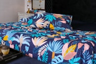 A bed is adorned with a vibrant and colorful floral-patterned bedding set. The design features an array of tropical leaves and abstract shapes in shades of blue, pink, yellow, and orange against a dark background. In the foreground, a decorative tray holds a small pot and a candle. The bed is situated in a well-decorated room with a vintage chest of drawers and a couple of elegant lamps.