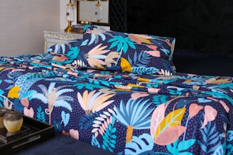 A bed is adorned with a vibrant and colorful floral-patterned bedding set. The design features an array of tropical leaves and abstract shapes in shades of blue, pink, yellow, and orange against a dark background. In the foreground, a decorative tray holds a small pot and a candle. The bed is situated in a well-decorated room with a vintage chest of drawers and a couple of elegant lamps.
