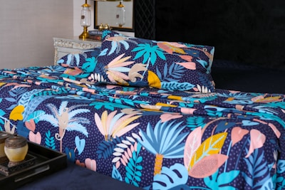 A bed is adorned with a vibrant and colorful floral-patterned bedding set. The design features an array of tropical leaves and abstract shapes in shades of blue, pink, yellow, and orange against a dark background. In the foreground, a decorative tray holds a small pot and a candle. The bed is situated in a well-decorated room with a vintage chest of drawers and a couple of elegant lamps.
