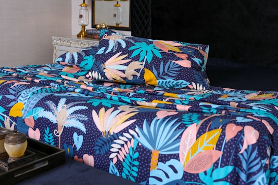 A bed is adorned with a vibrant and colorful floral-patterned bedding set. The design features an array of tropical leaves and abstract shapes in shades of blue, pink, yellow, and orange against a dark background. In the foreground, a decorative tray holds a small pot and a candle. The bed is situated in a well-decorated room with a vintage chest of drawers and a couple of elegant lamps.