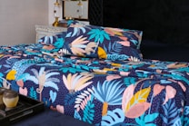 A bed is adorned with a vibrant and colorful floral-patterned bedding set. The design features an array of tropical leaves and abstract shapes in shades of blue, pink, yellow, and orange against a dark background. In the foreground, a decorative tray holds a small pot and a candle. The bed is situated in a well-decorated room with a vintage chest of drawers and a couple of elegant lamps.