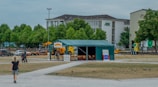 A green tent labeled 'Aboriginal Embassy' is set up in an open area surrounded by pathways and grassy fields. There are multiple signs displayed in front of the tent, some with text about Aboriginal democracy. A few people are near the tent and one person is walking toward it. In the background, several trees provide a green backdrop, and there is a large building with an 'AOK' logo. Parked vehicles are visible around the area.