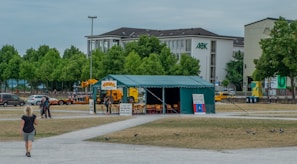 A green tent labeled 'Aboriginal Embassy' is set up in an open area surrounded by pathways and grassy fields. There are multiple signs displayed in front of the tent, some with text about Aboriginal democracy. A few people are near the tent and one person is walking toward it. In the background, several trees provide a green backdrop, and there is a large building with an 'AOK' logo. Parked vehicles are visible around the area.