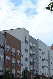 A series of modern multi-story apartment buildings with various exteriors including wood, brick, and siding. The buildings have numerous windows and small balconies. The sky above is partly cloudy with patches of blue visible. A few tree branches can be seen on the left.