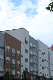 A series of modern multi-story apartment buildings with various exteriors including wood, brick, and siding. The buildings have numerous windows and small balconies. The sky above is partly cloudy with patches of blue visible. A few tree branches can be seen on the left.