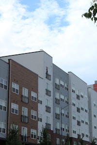 A series of modern multi-story apartment buildings with various exteriors including wood, brick, and siding. The buildings have numerous windows and small balconies. The sky above is partly cloudy with patches of blue visible. A few tree branches can be seen on the left.