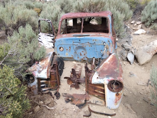 A heavily rusted and decaying old vehicle sits abandoned in a natural setting. The body is missing several parts, including the engine and wheels, leaving behind an empty shell. Surrounding it are shrubs and dry vegetation, indicating a possibly arid environment. The paint shows signs of peeling and wear, with exposed metal parts.