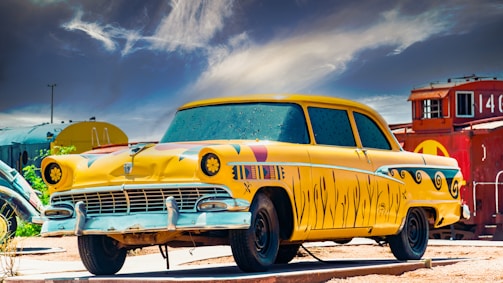 A vintage car is parked outdoors, painted in a bright yellow color with unique artistic patterns and black detailing. The car is set against a background of train cars and a dramatic sky with wispy clouds, creating a contrast between the colorful vehicle and the rustic surroundings.