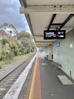 A train platform with an overhead digital display showing train information. There is a railway track running alongside the platform, which is bordered by greenery and trees. A row of tactile paving is visible on the platform, and there is a bench and a poster on the wall.