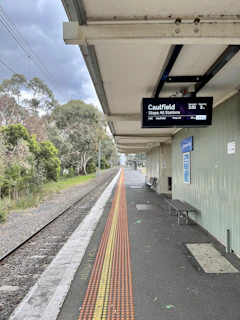 A train platform with an overhead digital display showing train information. There is a railway track running alongside the platform, which is bordered by greenery and trees. A row of tactile paving is visible on the platform, and there is a bench and a poster on the wall.