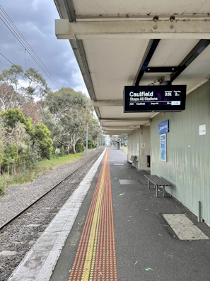 A train platform with an overhead digital display showing train information. There is a railway track running alongside the platform, which is bordered by greenery and trees. A row of tactile paving is visible on the platform, and there is a bench and a poster on the wall.
