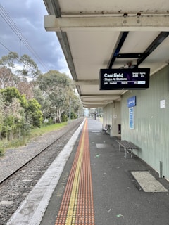 A train platform with an overhead digital display showing train information. There is a railway track running alongside the platform, which is bordered by greenery and trees. A row of tactile paving is visible on the platform, and there is a bench and a poster on the wall.