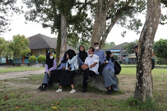 A group of people sit together under trees in an outdoor setting. They are wearing uniforms, some with masks, and appear to be engaged in conversation. The background shows buildings and a grassy area, indicating a school or campus environment.