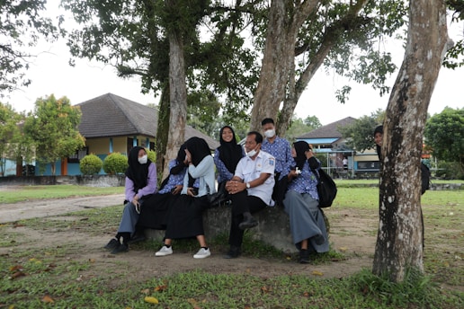 A group of people sit together under trees in an outdoor setting. They are wearing uniforms, some with masks, and appear to be engaged in conversation. The background shows buildings and a grassy area, indicating a school or campus environment.