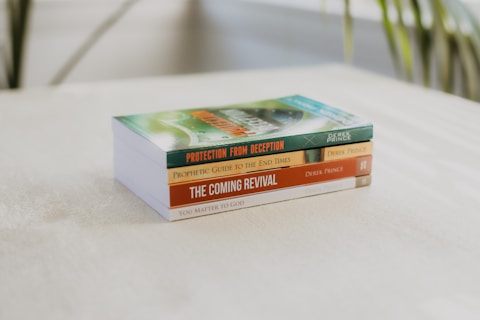 A stack of four books is neatly arranged on a light-colored surface with blurred greenery in the background. Each book is authored by Derek Prince, featuring titles like Protection from Deception and The Coming Revival.