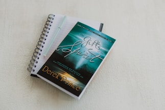 A spiral-bound notebook with lined pages lies open on a light-colored surface. A pen rests on top of the notebook. A book with a turquoise and green cover titled 'The Gifts of the Spirit' by Derek Prince is placed on top of the notebook.