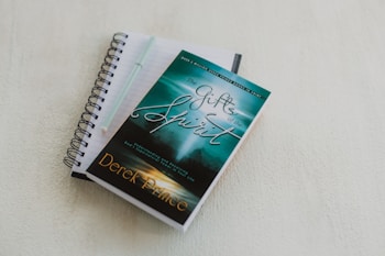 A spiral-bound notebook with lined pages lies open on a light-colored surface. A pen rests on top of the notebook. A book with a turquoise and green cover titled 'The Gifts of the Spirit' by Derek Prince is placed on top of the notebook.
