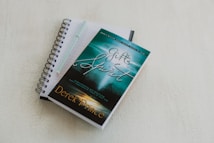 A spiral-bound notebook with lined pages lies open on a light-colored surface. A pen rests on top of the notebook. A book with a turquoise and green cover titled 'The Gifts of the Spirit' by Derek Prince is placed on top of the notebook.