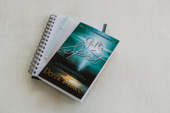 A spiral-bound notebook with lined pages lies open on a light-colored surface. A pen rests on top of the notebook. A book with a turquoise and green cover titled 'The Gifts of the Spirit' by Derek Prince is placed on top of the notebook.