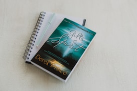 A spiral-bound notebook with lined pages lies open on a light-colored surface. A pen rests on top of the notebook. A book with a turquoise and green cover titled 'The Gifts of the Spirit' by Derek Prince is placed on top of the notebook.