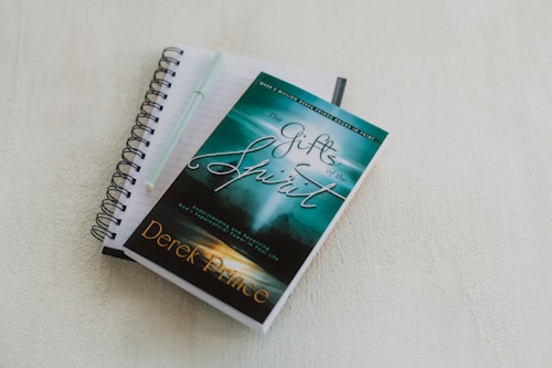 A spiral-bound notebook with lined pages lies open on a light-colored surface. A pen rests on top of the notebook. A book with a turquoise and green cover titled 'The Gifts of the Spirit' by Derek Prince is placed on top of the notebook.
